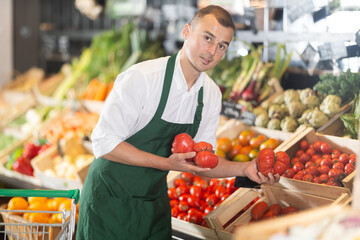 Friendly male seller in green apron enthusiastically offering fresh ripe tomatoes in organic...