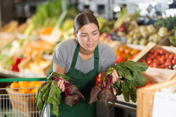 Smiling young saleswoman in green apron handling fresh beets, organizing organic produce on display...