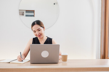 A woman is sitting at a desk with a laptop and a notebook. She is smiling and she is happy