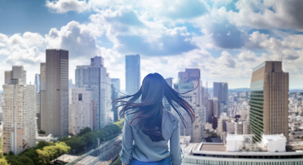 日本の街・都市・朝日を受けたオフィスビル群を前に佇み風を受けて髪をなびかせる会社員の女性の出勤　ビジネス・はじまり・決意・目標・挑戦のイメージ