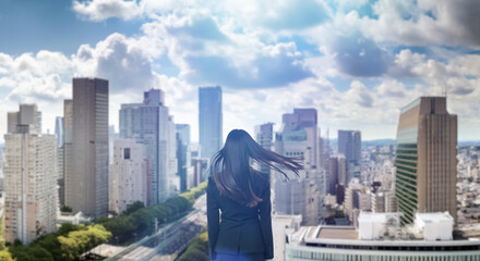 日本の街・都市・朝日を受けたオフィスビル群を前に佇み風を受けて髪をなびかせる会社員の女性の出勤　就職・ビジネス・はじまり・決意・目標・挑戦のイメージ