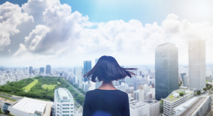 日本の街・都市・朝日を受けたオフィスビル群を前に佇み風を受けて髪をなびかせる会社員の女性の出勤　就職・ビジネス・はじまり・決意・目標・挑戦のイメージ