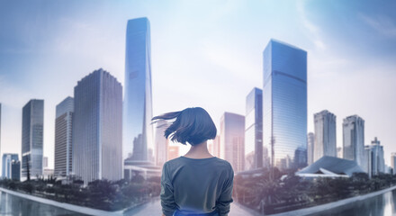 日本の街・都市・朝日を受けたオフィスビル群を前に佇み風を受けて髪をなびかせる会社員の女性の出勤　就職・ビジネス・はじまり・決意・目標・挑戦のイメージ