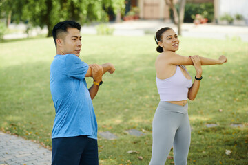 Man and woman in athletic wear stretching their arms together during an outdoor workout in the park