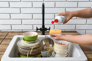 Woman washing dirty dishes with detergent in sink near white brick wall