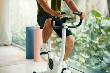 A man in workout clothes riding a stationary bike indoors, with plants visible through a large window