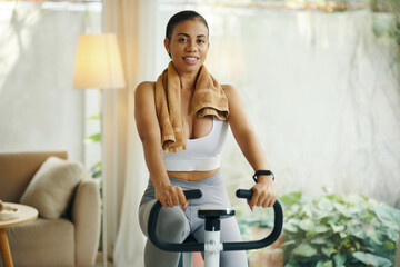 A woman in activewear resting on an exercise bike with a towel around her neck in a cozy living room