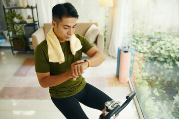 A man in workout attire checking his fitness tracker while exercising on a stationary bike indoors