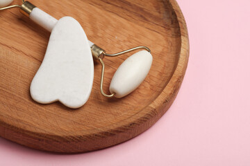 Massage roller and gua sha scraper for anti-aging facial massage on wooden plate, pink background.