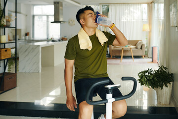 A man drinking water from a bottle after an intense workout session on a stationary bike at home