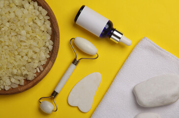 Jade facial roller and gua sha scraper, serum bottle, sea salt, towel on yellow background. Top...