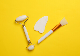 Jade facial roller and gua sha scraper, body butter spatula on yellow background. Beauty concept. Top view