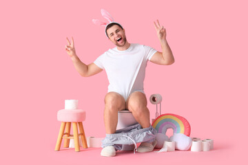 Young man in bunny ears showing victory gesture on toilet bowl against pink background