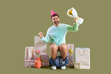 Young man in Birthday hat sitting on toilet bowl with gifts against green background