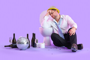 Drunk young businessman sitting near toilet bowl after party on lilac background