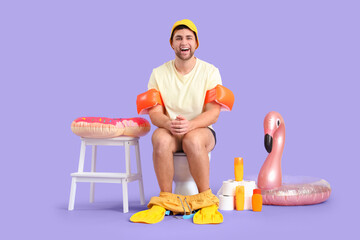 Male vacationer with beach accessories sitting on toilet bowl against lilac background