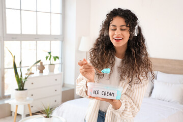 Beautiful young African-American woman in headphones with basket of ice cream in bedroom