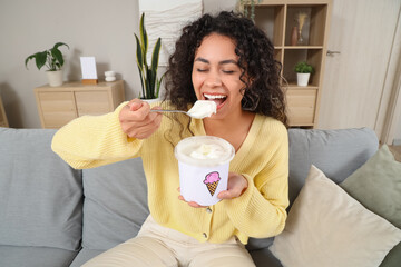 Beautiful young African-American woman eating ice cream and sitting on sofa at home