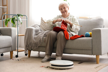 Happy senior woman knitting socks with robot vacuum cleaner at home