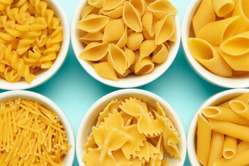 Bowls with different tasty uncooked pasta on blue background, closeup