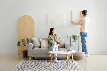 Young man hanging painting on light wall and his wife at home