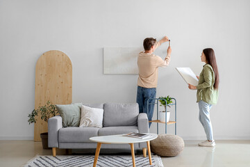 Young couple hanging paintings on light wall at home