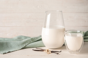 Glass and jug of fresh vanilla milk with sticks and flower on white wooden background
