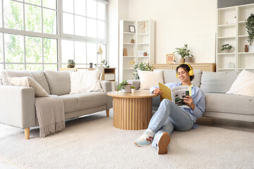 Young African-American woman in headphones reading magazine beside sofa at home