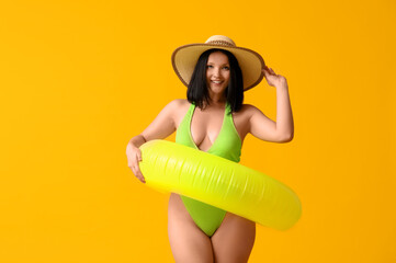 Beautiful young woman in stylish green swimsuit with inflatable ring on yellow background