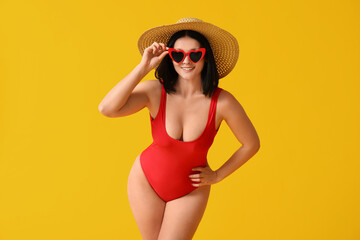 Beautiful young woman in stylish red swimsuit and sunglasses on yellow background