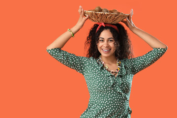 Beautiful young happy African-American woman with wicker plate full of kiwi on orange background