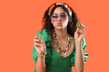 Beautiful young happy African-American woman with slices of kiwi blowing kiss on orange background