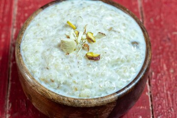 Closeup of Cracked Wheat Kheer or Payesh Garnished with Cashew, Almond, Pistachio in a Wooden Bowl Isolated on Red Wooden Background with Copy Space, Also Known as Ksheeram, Milk Pudding