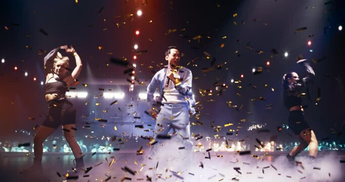 Energetic Pop Artist in Casual Costume Performing on Stage with Two Young Female Dancers in Alternative Clothes. Excited Audience Illuminated by Vibrant Light Show. Glittering Confetti Falling Down