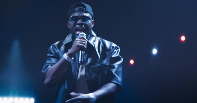 RnB Musician Gives a Breathtaking Performance Under a Canopy of Lights. Stunning Visual Effects Enrich the Hip Hop Concert. Zoom In Portrait of a Black Man in Leather Jacket, Shades and a Cap