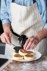 Hands of a female chef placing freshly cooked syrniki on a plate, garnishing with sour cream and mint. Concept of home cooking, traditional recipes, and culinary presentation.