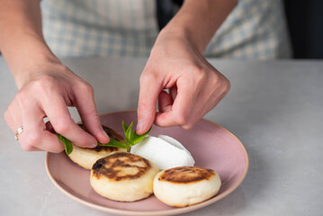 Hands of a female chef placing freshly cooked syrniki on a plate, garnishing with sour cream and mint. Concept of home cooking, traditional recipes, and culinary presentation.