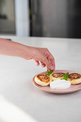 Hands of a female chef placing freshly cooked syrniki on a plate, garnishing with sour cream and mint. Concept of home cooking, traditional recipes, and culinary presentation.