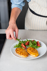 Fish cutlets being placed on a plate and garnished with arugula salad and cherry tomatoes. Concept of home cooking, traditional healthy food recipes, and culinary presentation.