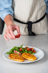 Fish cutlets being placed on a plate and garnished with arugula salad and cherry tomatoes. Concept of home cooking, traditional healthy food recipes, and culinary presentation.