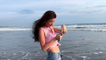 A beautiful girl takes pictures of the ocean on her smartphone. A woman walks along the coast of a popular beach and shoots a video of foaming sea waves at sunset for social media. . High quality 4k