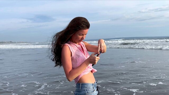 A beautiful girl takes pictures of the ocean on her smartphone. A woman walks along the coast of a popular beach and shoots a video of foaming sea waves at sunset for social media. . High quality 4k 