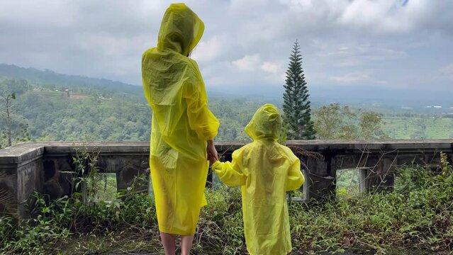 A rear view of a mother and child standing on the terrace of hotel in yellow raincoats and looking at the picturesque views of nature. Tourists explore the abandoned hotel Pondok Indah Bedugul In Bali