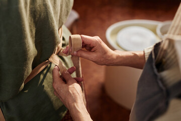 Close up of unrecognizable senior woman helping client tying apron during pottery class in art...