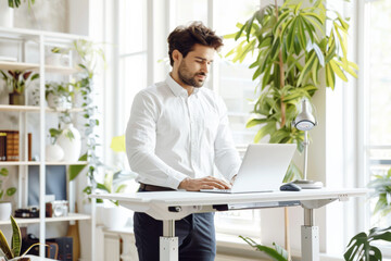 Man working on a laptop at a standing desk in a modern office with plants.