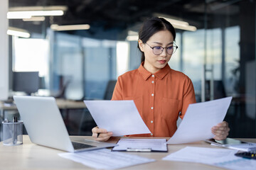 Asian financial woman working inside office with documents, papers behind paperwork, serious businesswoman looking financial reports at workplace , confident successful female accountant.