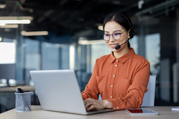 Asian woman with headset phone smiling working inside office with laptop, online customer support...