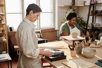Side view portrait of senior woman setting up vinyl record for music ambiance during pottery class copy space