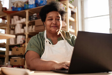 Warm toned portrait of African American senior woman using laptop in art studio lit by sunlight...