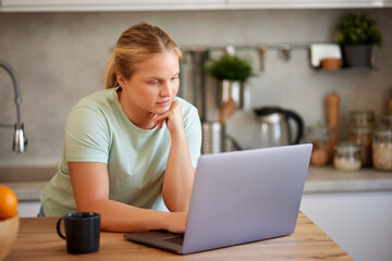 Young woman wearing casual clothes using laptop and drinking coffee in the kitchen